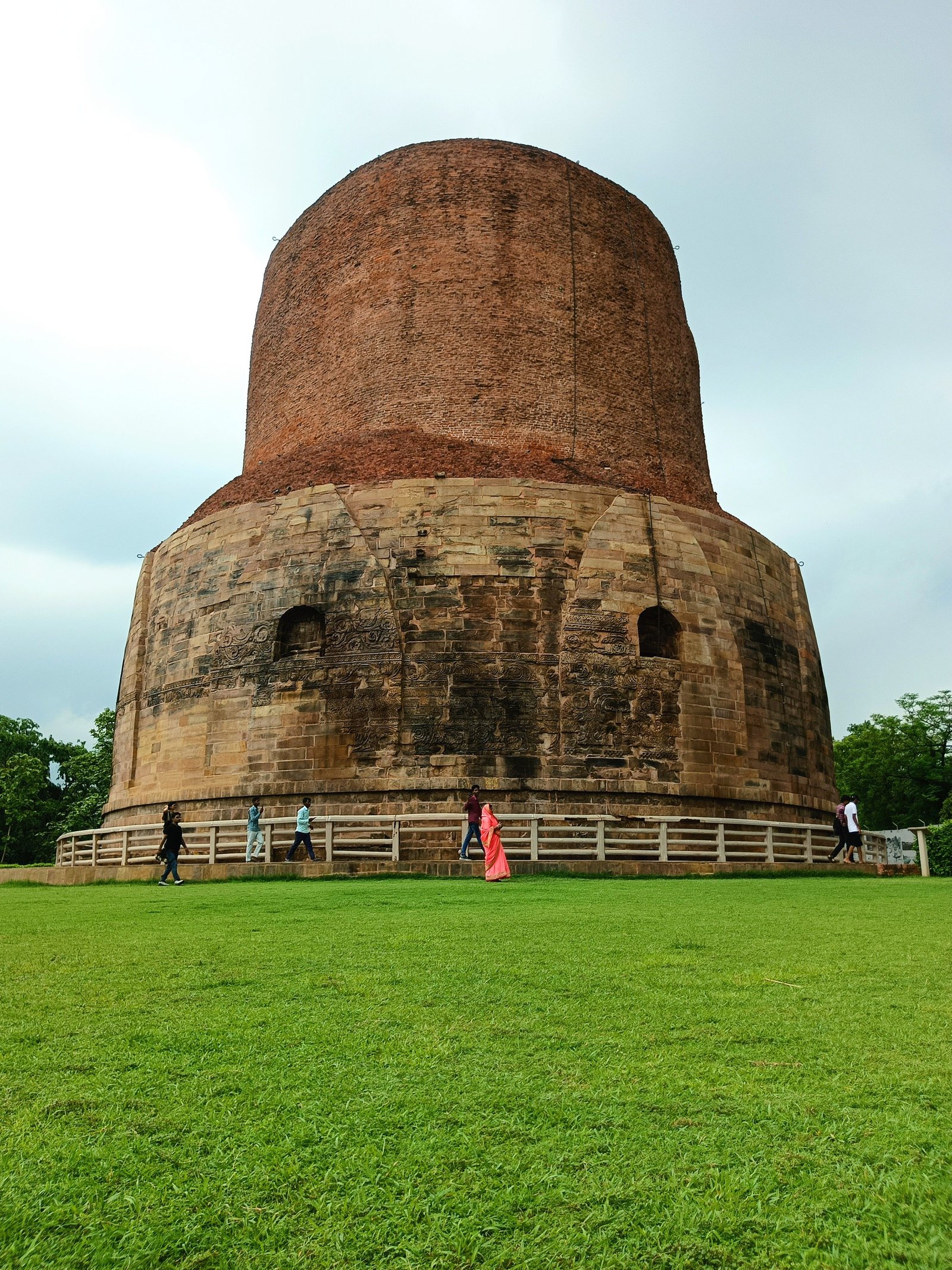 Dhamek Stupa at Sarnath, with its tall brick cylinder, carved stone base and visitors beside the railing.